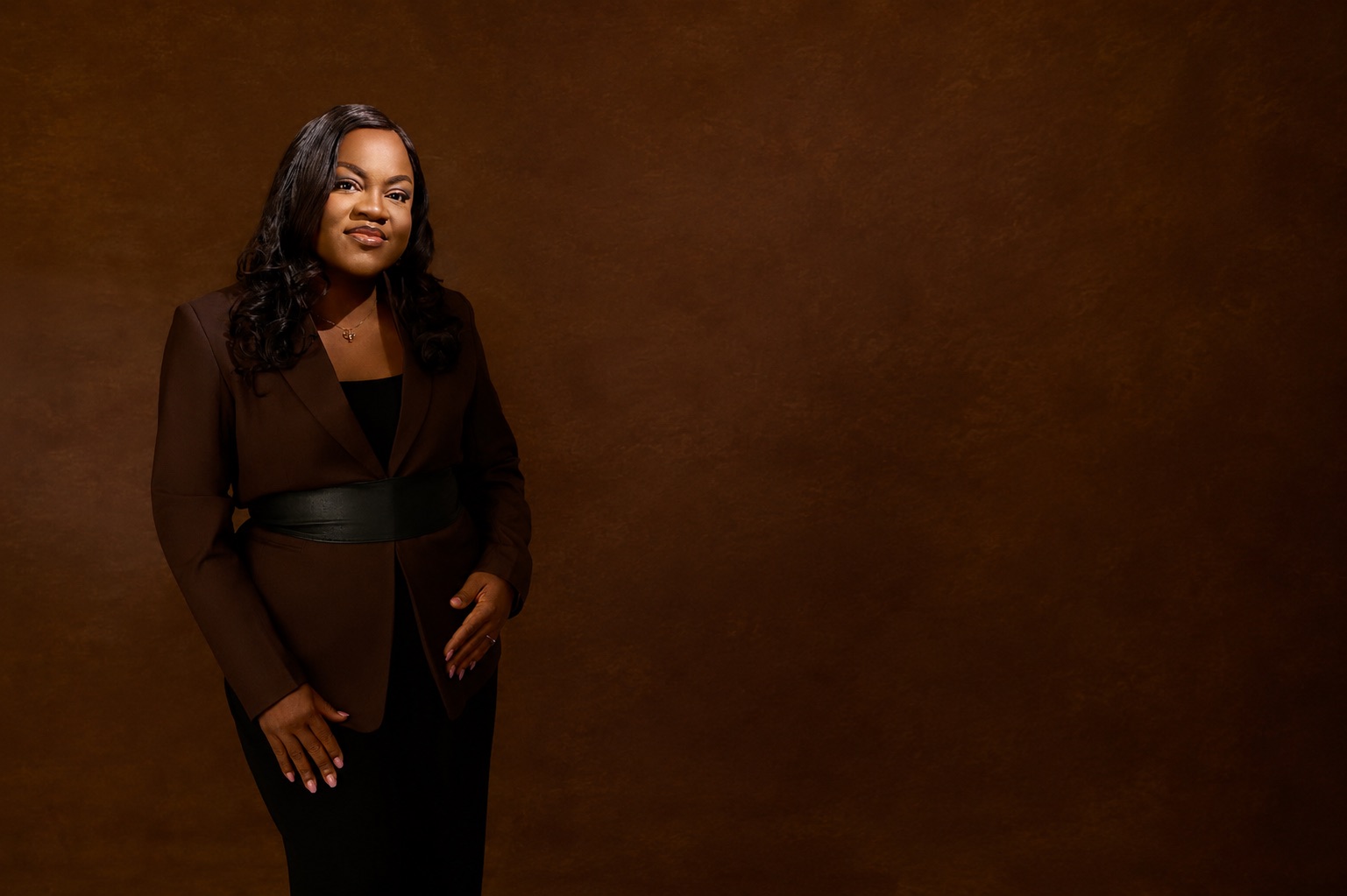 Busayo Disu standing in a brown suit against a warm brown studio backdrop