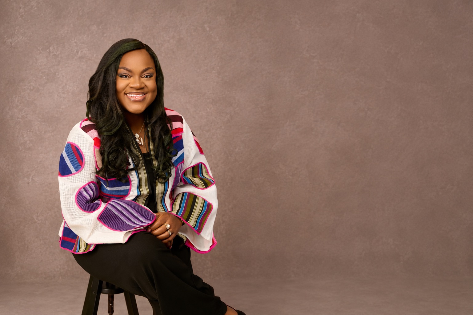 Busayo Disu seated on a stool in a colourful striped African print jacket, smiling