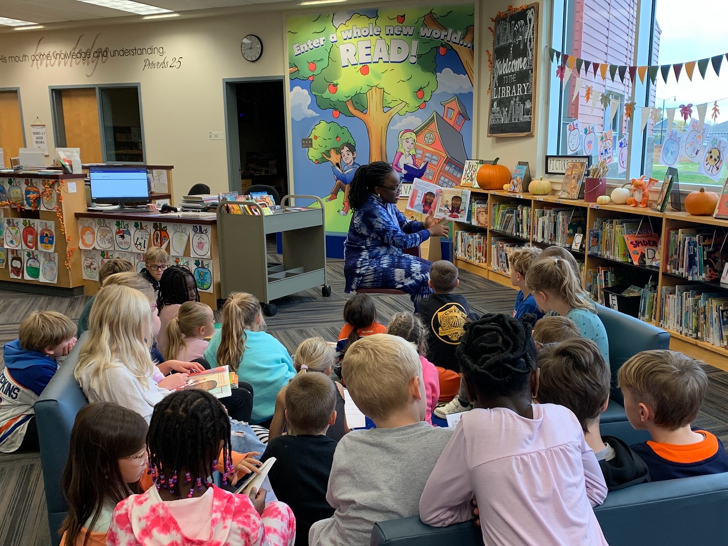 Busayo Disu reading a picture book aloud to a school library full of children seated on the carpet