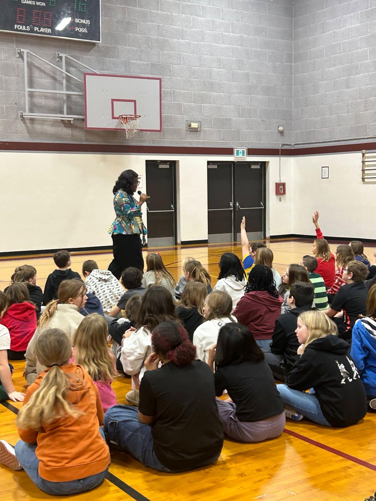 A room of children with their hands up to answer Busayo Disu at a school assembly