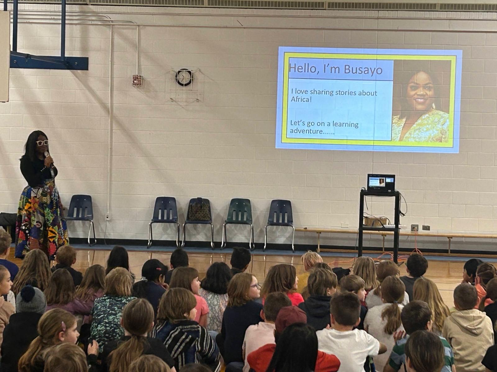 Busayo Disu opening a school assembly, her &ldquo;Hello, I&rsquo;m Busayo&rdquo; slide on the screen and children seated in front of her