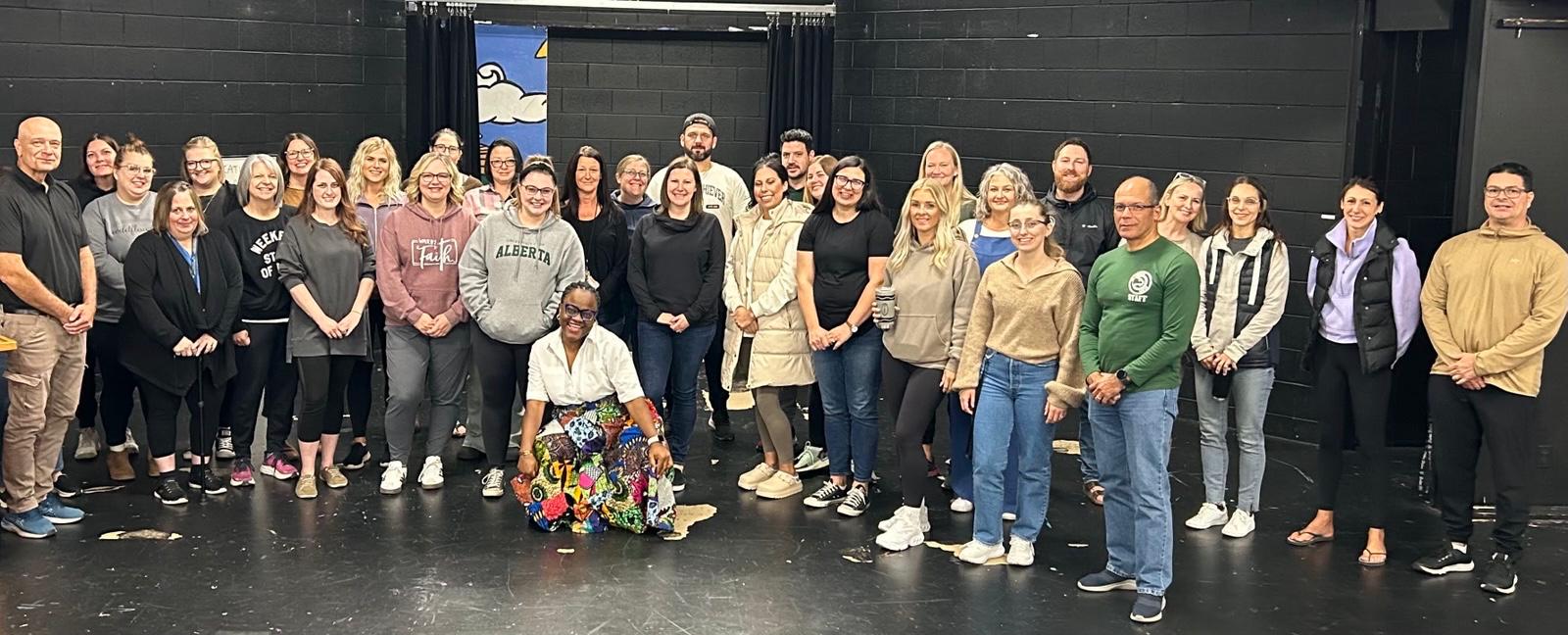 A school staff group gathered on a theatre stage, with Busayo Disu kneeling at the front of them