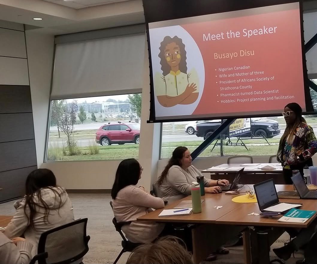 Busayo Disu speaking beside a slide reading Meet the Speaker, to a room seated around a boardroom table